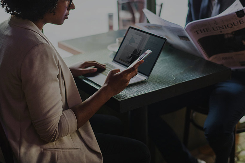 black woman sitting at a small table working on a laptop and reading her phone opposite a man reading newspaper